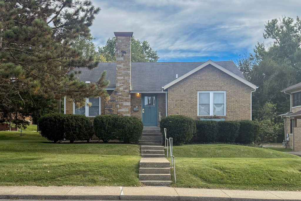 Front of 1314 E Atwater Ave in Bloomington, IN, a one-story tan brick house with a tall stone chimney, a gabled front wing and a teal front door reached by concrete steps up a wide lawn