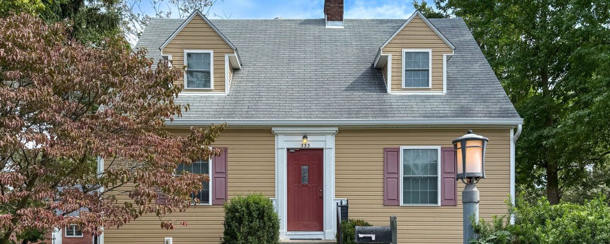 Front of 333 S Eagleson Ave in Bloomington, IN, a Cape Cod-style house with tan siding, two dormers, a red front door and burgundy shutters