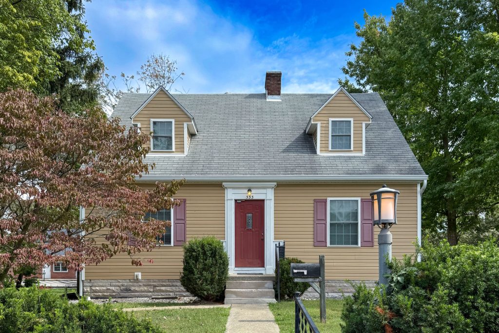 Front of 333 S Eagleson Ave in Bloomington, IN, a Cape Cod-style house with tan siding, two dormers, a red front door and burgundy shutters